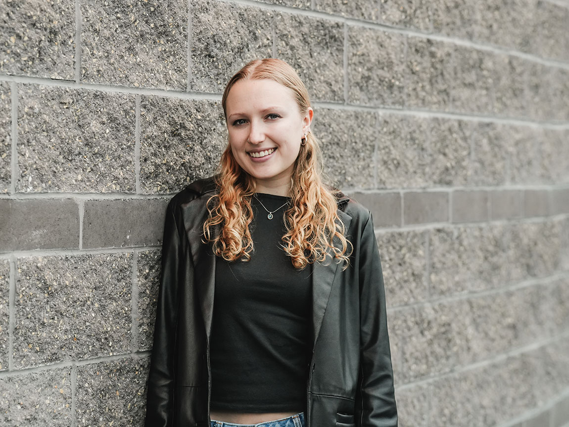 A college student wearing a black shirt and jacket smiles with a gray brick wall behind her